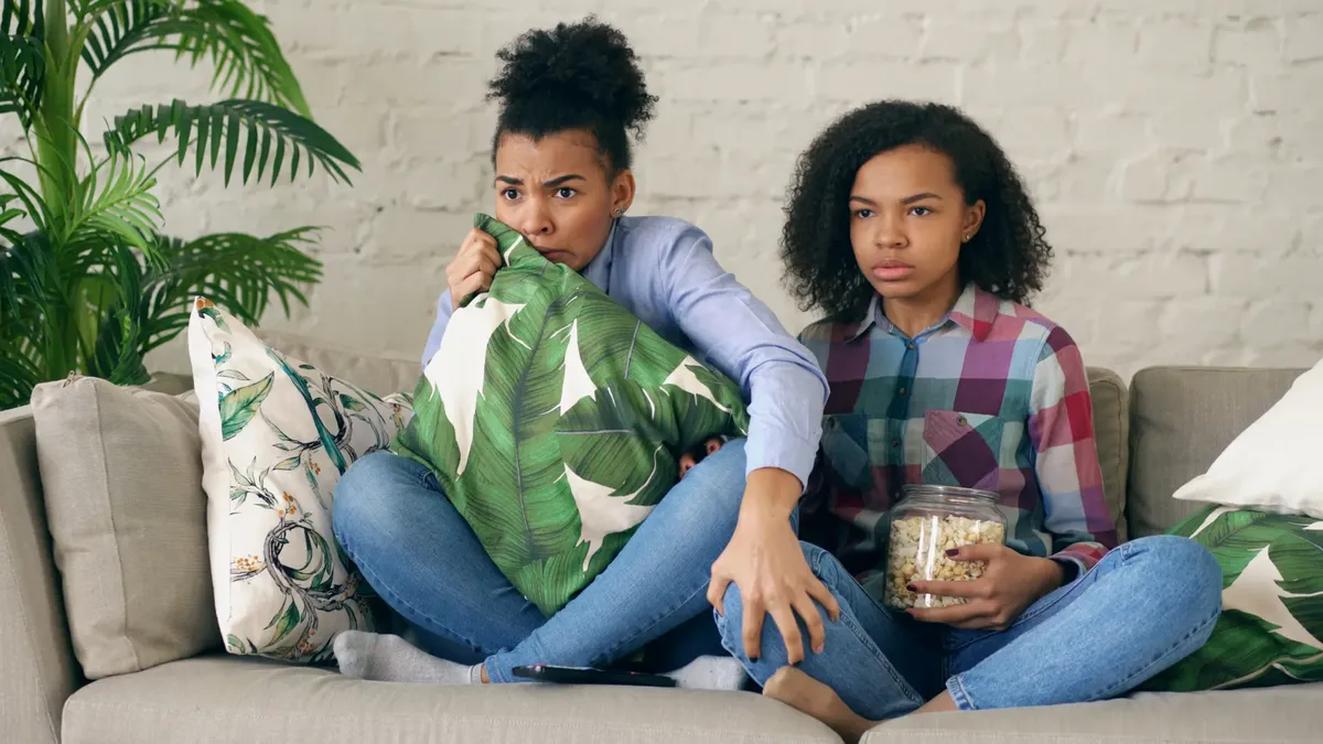 Two teenagers watching a movie at home, one holding a pillow with a jar of popcorn beside them.